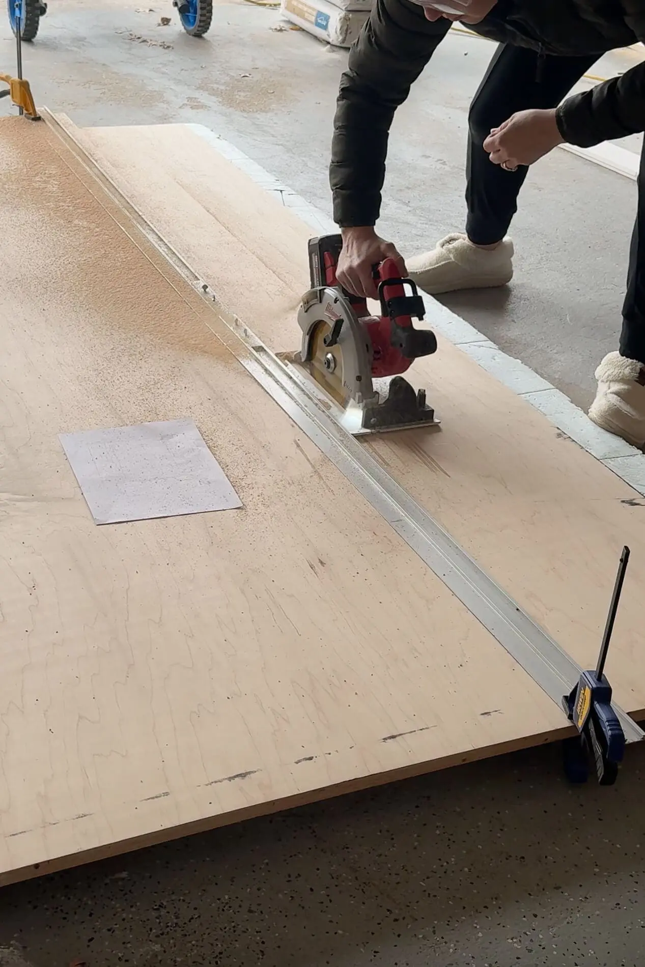 A DIYer in a green puffer jacket and fuzzy slippers using a red and black circular saw to cut a large sheet of plywood. A metal guide rail and clamps help ensure a straight cut. Sawdust is visible spraying from the cut as the person carefully follows the line.